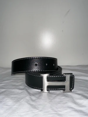 Svart skärp med H-spänne - Snyggt svart bälte i skinn med kontrasterande vita sömmar och ett stort H-format spänne i silverfärg. Perfekt accessoar för att lyfta din outfit med en clean och stilren look.