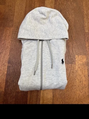 Grå hoodie från Ralph Lauren - Snygg ljusgrå hoodie från Ralph Lauren med dragkedja framtill och justerbara snören i huvan. Klassisk svart logga broderad på bröstet. Perfekt för en avslappnad och stilren look.