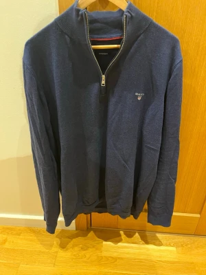 Marinblå stickad halvzip från Gant - Stickad marinblå tröja från Gant med halv dragkedja och hög krage. Tröjan har ribbade muddar och diskret Gant-logga broderad på bröstet. Perfekt för lager på lager och enkel att styla till olika looks.