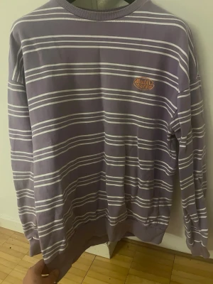 Lila randig tröja från Multiverse - Säljer en lila långärmad tröja från Multiverse med vita horisontella ränder och broderad logga i orange på bröstet. Tröjan har ribbade muddar vid ärmslut och nederkant. Perfekt för en avslappnad och trendig stil.