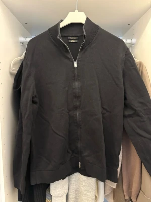 Svart zip-cardigan från Massimo Dutti - Svart cardiganjacka från Massimo Dutti i 100% bomull. Jackan har en hög krage, hel dragkedja och ribbade detaljer vid krage, ärmslut och nederkant. 