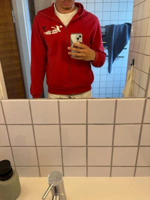 Röd hoodie från Adidas med dragkedja - Röd hoodie från Adidas med vit logga och siffran 7 på bröstet. Den har dragkedja framtill, två fickor och en huva. Materialet är mjukt och bekvämt, perfekt för att chilla eller hänga med kompisar.