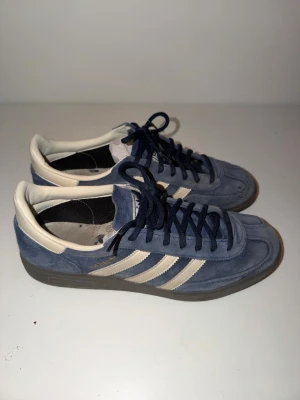 Adidas blå sneakers med vita ränder - Snygga Adidas sneakers i blå mocka med klassiska vita ränder på sidorna och mörkblå snörning. Skorna har en låg profil och gummisula, perfekt för en avslappnad streetstyle-look. Ikonisk design med retrovibbar och bekväm passform.