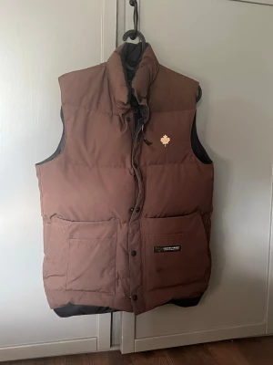 Brun dunväst från Canada Goose - Snygg brun dunväst från Canada Goose med hög krage och två stora fickor framtill. Västen har en ikonisk lönnlöv-logga på bröstet och stängs med både dragkedja och knappar. Perfekt för lager-på-lager och håller dig varm under kyliga dagar.