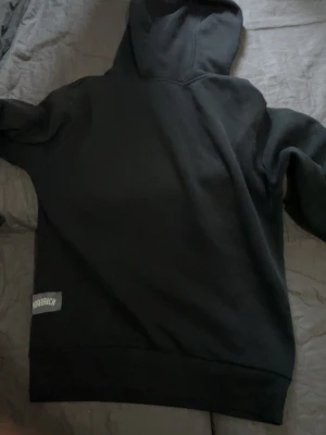 Svart hoodie från Hoodrich - Snygg svart hoodie från Hoodrich med klassisk huva och ribbade muddar. Hoodien har en diskret patch med Hoodrich-loggan på ärmen. Perfekt för en avslappnad och street-inspirerad stil.