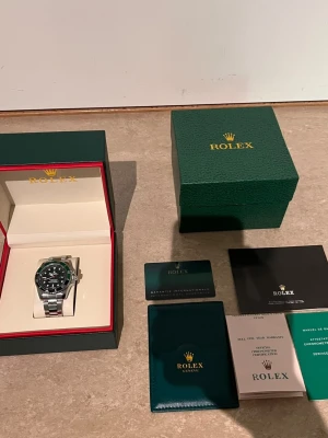 Role Submariner med grön bezel - Snygg Role  Submariner med grön bezel och svart urtavla. Klockan har ett robust armband i silverfärgat stål och klassisk rund form. Levereras med originalbox, certifikat och manualer. Perfekt för dig som gillar exklusiva accessoarer.