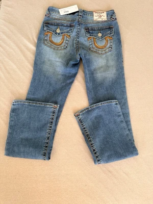 True Religion bootcut jeans blå - Snygga blå bootcut jeans från True Religion med klassiska kontrastsömmar och detaljerade bakfickor med lock och knappar. Jeansen har en lätt tvättad look och är tillverkade i denim med orangea sömmar och silverfärgade knappar.