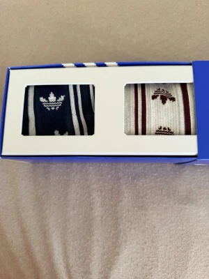Adidas randiga strumpor 2-pack - Två par Adidas strumpor i klassisk retrostil. Ett par är marinblå med vita ränder och Adidas-logga, det andra är vita med vinröda detaljer och logga. Båda paren har ribbade muddar och är tillverkade i mjuk bomullsblandning. Perfekta för dig som gillar sportig streetwear.