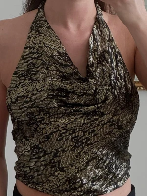 Y2K guldig halterneck topp - Superfin halterneck topp i svart/guld med skimrande material och draperad urringning. Väldigt snygg till fest, middag eller utekväll och ger en glam/Y2K vibe. Passar perfekt med både jeans, kostymbyxor eller kjol