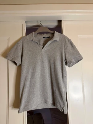 Grå pikétröja från Massimo Dutti - Snygg grå pikétröja från JLindberg med klassisk krage och korta ärmar. Tillverkad i mjuk bomull som känns skön mot huden. Perfekt för en clean och stilren look.