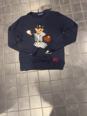 Mörkblå stickad tröja Polo Ralph Lauren - Mörkblå stickad tröja från Polo Ralph Lauren med en ikonisk nalle i basebollkläder på framsidan. Tröjan har rund hals, ribbstickade muddar och RL-broderi nertill. Perfekt för dig som gillar sportiga och lekfulla detaljer.