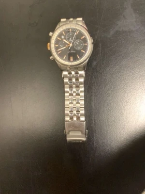 Silverfärgad Seiko kronografklocka - Stilren Seiko kronografklocka i silver med svart urtavla och detaljer i orange. Klockan har länkarmband i metall och flera små urtavlor för extra funktioner. Perfekt accessoar för dig som gillar klassisk och sportig stil.
