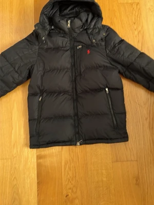 Polo Ralph Lauren jacka svart - Svart pufferjacka från Ralph Lauren med huva och dragkedja framtill. Jackan har två sidofickor med dragkedja och en bröstficka med dragkedja. Klassisk röd logga på bröstet. Perfekt för kyliga dagar. Jackan är äkta direkt från Ralph Lauren