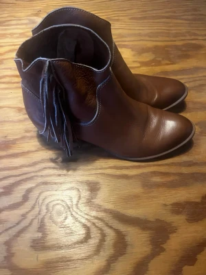  Boots med fransar - Bruna boots i läder med fransar på sidan och kontrastsömmar. Bootsen har en låg klack, samt dragkedja på insidan för enkel påtagning. 