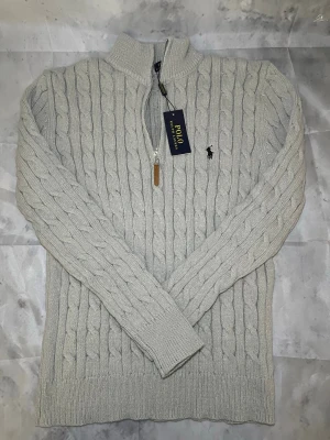 Ljusgrå kabelstickad tröja Polo Ralph Lauren - Snygg ljusgrå kabelstickad tröja från Polo Ralph Lauren med halv dragkedja och hög krage. Tröjan har det klassiska broderade logotypen på bröstet och ribbade muddar vid ärmslut och nederkant. Perfekt för lager-på-lager-stil.