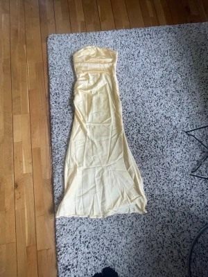 Ljusgul axelbandslös långklänning - Elegant ljusgul långklänning i ett glansigt material med axelbandslös design. Klänningen har en figurnära passform. (Inte struken) jätte fin men säljer då den va för stor. 