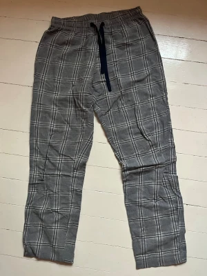 Rutiga grå pyjamasbyxor Ellos - Mysiga pyjamasbyxor i grått med vitt rutmönster och svart dragsko i midjan från Ellos. Byxorna har avslappnad passform och resår i midjan för extra komfort. Perfekta för chillkvällar hemma eller när du bara vill ta det lugnt.