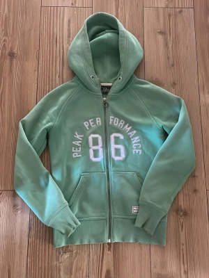 Grön hoodie Peak Performance 86 - Snygg grön hoodie från Peak Performance med dragkedja och stor huva. Framtill står det 'Peak Performance 86' i vita bokstäver. Hoodien har två fickor och ribbade muddar vid ärmslut och nederkant. Perfekt för en avslappnad och sportig stil.