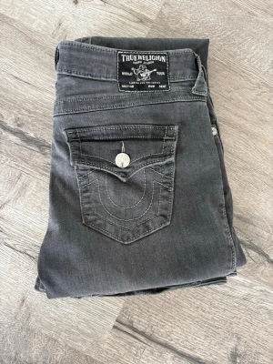 Gråa True Religion jeans - Säljer ett par gråa jeans från True Religion med ikonisk fickdesign och kontrastsömmar.  silverfärgade knappar med logga. Som man ser på andra bilden har sömmen lossnat men efter som det inte är en sånn söm som håller ihop jeansen så är det inget som påverkar kvaliten men med en nål så är det fixat på noll tid!❤️