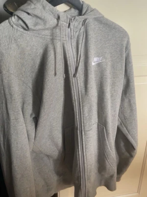 Grå zip hoodie från Nike - Klassisk grå hoodie från Nike med dragkedja framtill, justerbar huva med snören och fickor på sidorna. Tillverkad i mjukt sweatshirtmaterial och har Nike-logga broderad i vitt på bröstet. Perfekt för en avslappnad och sportig stil. Ny pris 1000kr.