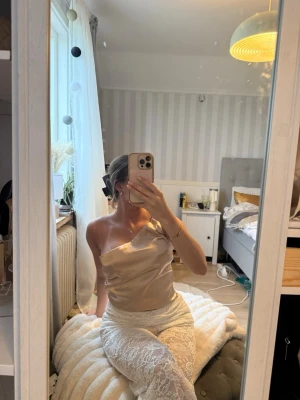 Champagnefärgad satin one shoulder topp - Snygg och trendig one shoulder topp i champagnefärgad satin. Toppen har en slät, glansig yta och sitter figurnära. Perfekt för dig som vill sticka ut med en elegant och modern look.