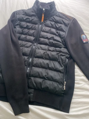 Svart pufferjacka från Parajumpers - Säljer en svart pufferjacka från Parajumpers med quiltad front och ryggparti, samt mjuka stickade ärmar. Jackan har hög krage, dragkedja framtill och två sidofickor med dragkedja. Märkets patch syns på ena ärmen. Perfekt för kalla dagar.