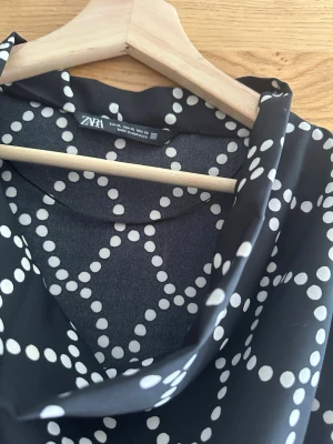 Svart prickig blus från Zara - Snygg svart blus från Zara med vita prickar i ett grafiskt mönster. Blusen har en lös passform, draperad krage och långa ärmar. Materialet är glansigt och känns lyxigt, perfekt för att lyfta vilken outfit som helst.