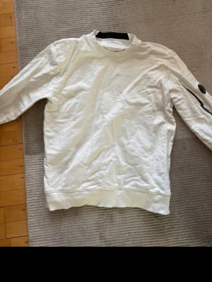 Vit sweatshirt från C.P. Company - Vit sweatshirt från C.P. Company med rund halsringning och ribbade muddar. Tröjan har en ikonisk svart linsdetalj på ärmen samt en dragkedjeficka. Perfekt för en clean och modern streetwear-look.