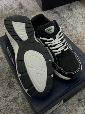 Svarta Dior sneakers  - Snygga svarta Dior sneakers med vita och grå detaljer samt vita skosnören. Ovandelen är i mesh och mocka med coola kontrastpaneler och en chunky vit sula. Perfekta för dig som vill ha en exklusiv och trendig look.