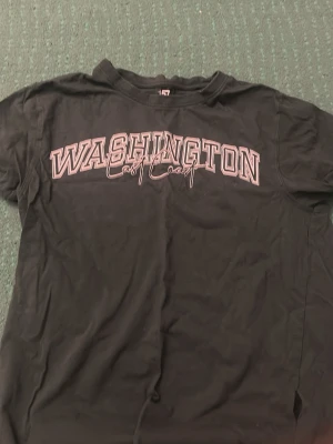Svart t-shirt med Washington-tryck - Cool svart t-shirt med stort 'WASHINGTON East Coast'-tryck över bröstet. Klassisk passform och rund halsringning. Perfekt för dig som gillar college-stil och streetwear.