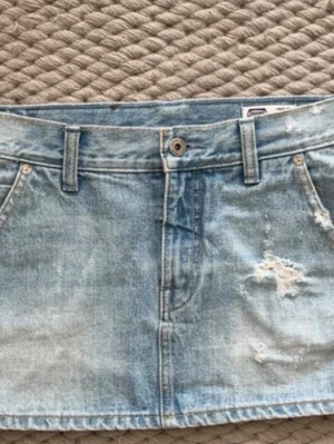 Ljusblå jeanskjol med slitningar - Trendig ljusblå jeanskjol med slitna detaljer och klassiska fickor fram. Kjolen har bälteshällor och knappstängning i midjan. Perfekt för dig som gillar en avslappnad och cool stil.