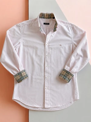 Ljusrosa skjorta från Burberry - Snygg ljusrosa skjorta från Burberry med klassiskt rutigt mönster på insidan av krage och ärmslut. Skjortan har knappar framtill och lång ärm. Perfekt för dig som gillar stilrena detaljer och ikonisk design. Hellängd - 73 cm Bredd - 54cm Helärmlängd - 62,5cm