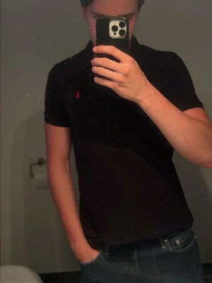 Svart pikétröja från Ralph Lauren - Klassisk svart pikétröja från Ralph Lauren med korta ärmar och den ikoniska röda logon broderad på bröstet. Tröjan har krage och knappar vid halsen. Väldigt clean piké passar bra till allt👏