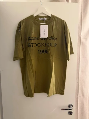 Olivgrön Acne Studios t-shirt herr M - Cool olivgrön t-shirt från Acne Studios med svart tryck på bröstet där det står 'Acne Studios STOCKHOLM 1996'. Klassisk rund hals och avslappnad passform. Perfekt för dig som gillar stilrena och trendiga plagg.
