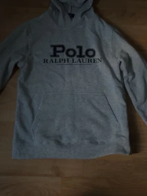Grå hoodie från Polo Ralph Lauren - Snygg grå hoodie från Polo Ralph Lauren med stor svart logga på bröstet. Klassisk modell med huva och magficka, perfekt för en avslappnad och trendig stil. Mjuk bomullsblandning som känns skön mot huden.