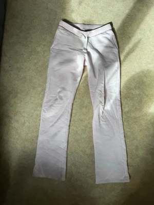 Ljusrosa mjukisbyxor med dragsko - Säljer ett par ljusrosa mjukisbyxor med vit dragsko i midjan. Byxorna har utsvängda ben och är tillverkade i ett mjukt och bekvämt material, perfekt för en avslappnad stil. Midjan är elastisk och byxorna har en enkel design utan tryck eller logga.