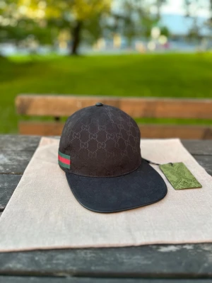 Svart keps från Gg med GG-mönster - Snygg svart keps från Gg med klassiskt GG-monogram över hela kepsen. Kepsen har en böjd skärm och detaljer i rött och grönt på sidan. Perfekt accessoar för att lyfta din streetwear-look.