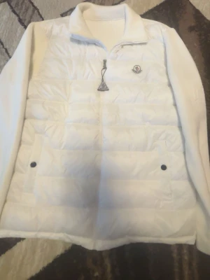 Vit cardiganjacka från Moncler  - Snygg vit dunväst från Moncler med quiltad framsida, dragkedja och två knäppbara fickor. Västen har ribbstickade ärmar och baksida i mjukt material samt Moncler-logga på bröstet. Perfekt för lager på lager och en clean look.