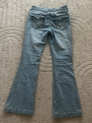 Ljusblå utsvängda jeans från H&M - Säljer ett par ljusblå jeans från H&M med utsvängda ben och justerbar midja. Jeansen har två bakfickor med lock och knappdetaljer samt en mid waist-passform. Sitter perfekt på mig som är 163 cm lång🩵