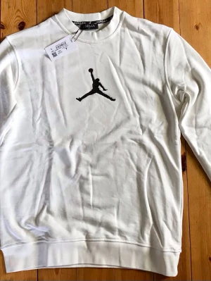 Vit Jordan sweatshirt med logga - Säljer en vit sweatshirt från Jordan med den klassiska svarta Jumpman-loggan broderad på bröstet. Tröjan har rund hals, långa ärmar och ribbade muddar. Perfekt för dig som gillar sportig och stilren design.