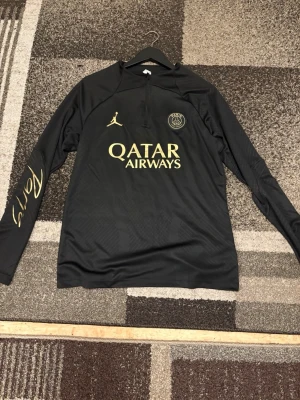 Svart PSG träningströja Jordan - Snygg svart långärmad träningströja från PSG x Jordan med guldfärgade detaljer och Qatar Airways-tryck på bröstet. Halvlång dragkedja framtill och diskret PSG-logga. Tillverkad i lätt och ventilerande material, perfekt för fotbollsträning.