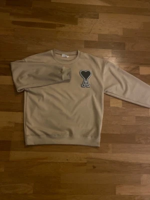 Beige sweatshirt med hjärta från Ami - Snygg beige sweatshirt från Ami med deras ikoniska svarta hjärta och A-logga broderad på bröstet. Tröjan har rund halsringning och ribbade muddar. Perfekt för en avslappnad och trendig look.