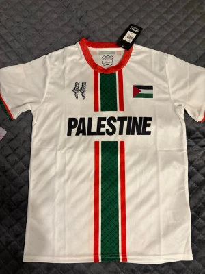 Vit Palestina fotbollströja S - Snygg vit fotbollströja från PC Palestina med gröna och röda ränder, svart text 'PALESTINE' och palestinska flaggan på bröstet. Röd krage och detaljer på ärmarna. Tillverkad i lätt och ventilerande material, perfekt för träning eller match.