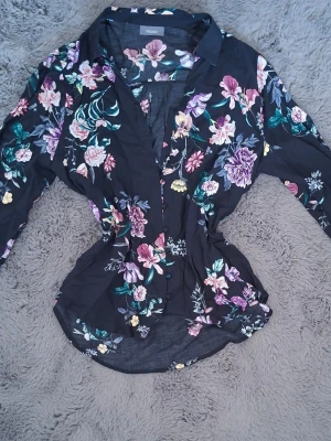Svart blommig blus med krage - Snygg svart blus från Vero Moda med färgglatt blommönster i lila, rosa, grönt och gult. Blusen har krage, knäppning framtill och långa ärmar. Perfekt för dig som gillar en feminin och trendig stil. Materialet känns lätt och luftigt.