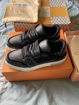 Louis Vuitton svarta sneakers - Snygga svarta Louis Vuitton sneakers i lågt utförande med ovandel i läder och detaljer i mocka. Skorna har vita sulor och klassisk LV-logga på sidan. Perfekta för dig som vill ha en exklusiv och stilren look.