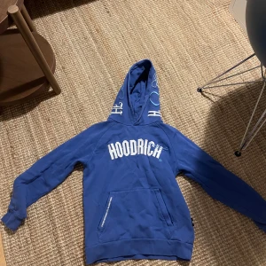 Blå hoodie från Hoodrich - Säljer en snygg blå hoodie från Hoodrich i storlek S. Den har en stor vit logga på bröstet och coola detaljer på huvan. Perfekt för en avslappnad stil och passar bra till både höst och vår. Den har en praktisk magficka och är i mycket bra skick.
