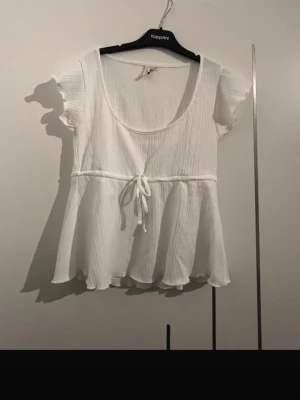 Vit blus med knyt och volang - Söt vit blus med korta ärmar, rundad urringning och knyt i midjan. Toppen har volangkant nedtill och är tillverkad i ett lätt, krinklat tyg som ger en avslappnad känsla. Perfekt för dig som gillar en feminin och luftig stil.
