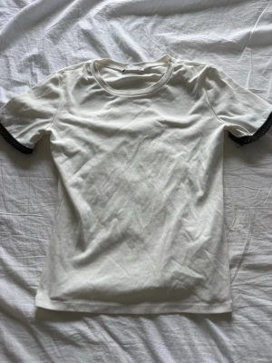 Vit t-shirt med spetsdetalj - Snygg vit t-shirt i bomull med korta ärmar och svart spetsdetalj vid ärmsluten. Enkel och stilren modell med rund halsringning, perfekt för en clean look.