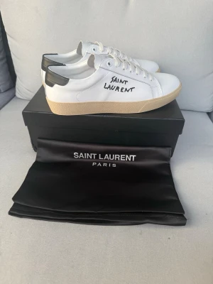 Saint Laurent vita sneakers - Snygga vita sneakers från Saint Laurent med svart detalj på hälen och handskriven logga på sidan. Skorna har en beige sula och klassisk låg profil. Tillverkade i läder med vita skosnören och stilren design som passar till det mesta.