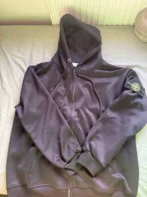 Svart hoodie från Stone Island - Säljer en svart hoodie från stone island med dragkedja och klassisk logotyp-patch på vänster ärm. Hoodien har ribbade muddar, fickor framtill och justerbar huva med snören. Perfekt för dig som gillar streetwear och vill ha en stilren look.